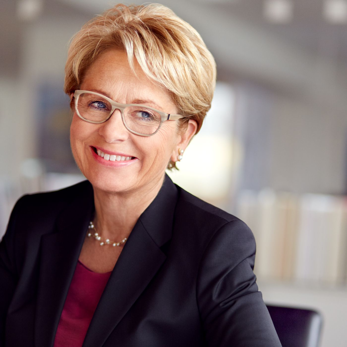 CEO-Portrait einer lächelnden Frau mit heller Brille und Perlenkette, entspannt zur Seite blickend vor unscharfer Bücherwand